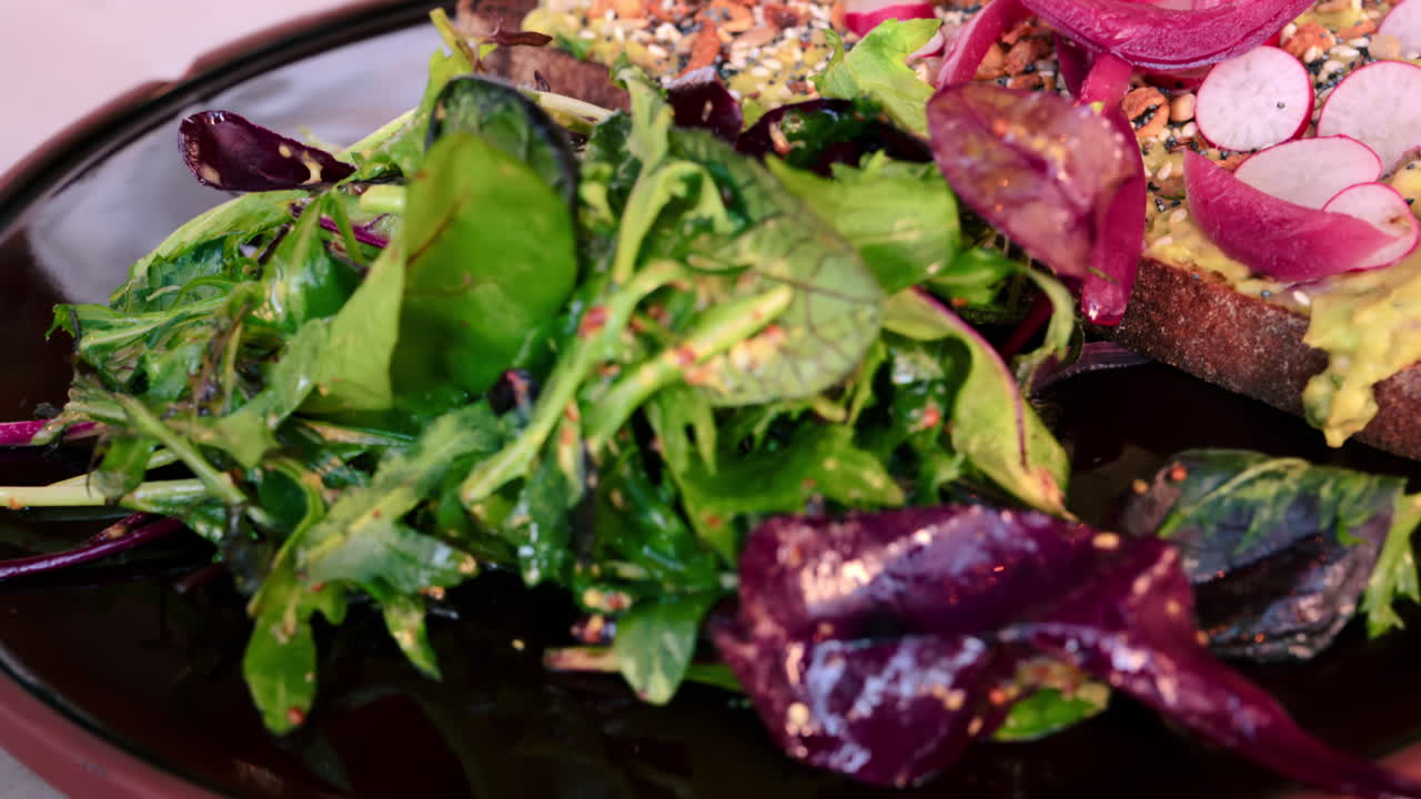 Close up of an avocado toast with seeds, radish, red onion and a side salad on a black plate