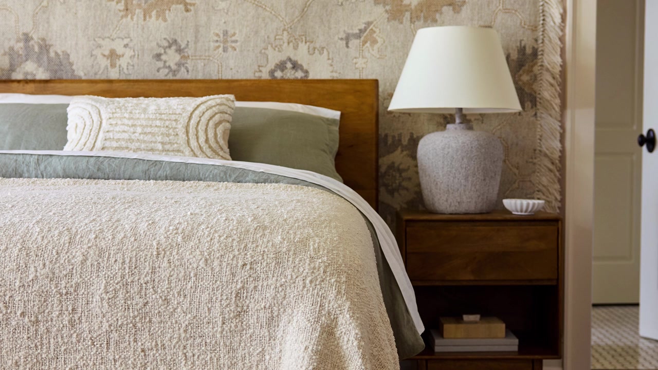 Lento zoom indietro di un angolo letto in stile rustico con biancheria da letto in lino a strati, una struttura in legno, una lampada grigia e crema, un comodino e carta da parati decorativa
