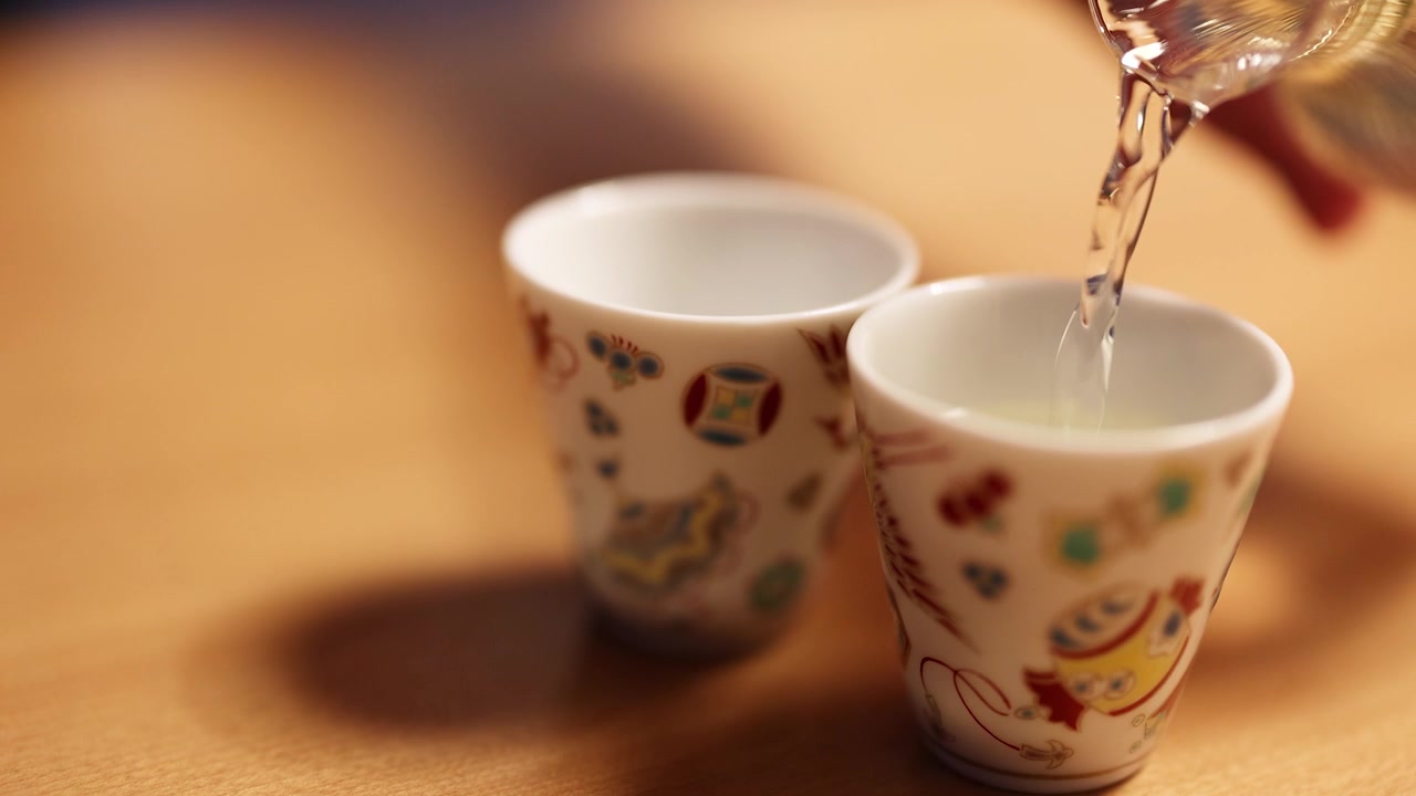 Sake limpido viene versato da una caraffa di vetro in tazze di ceramica decorate su un tavolo di legno.