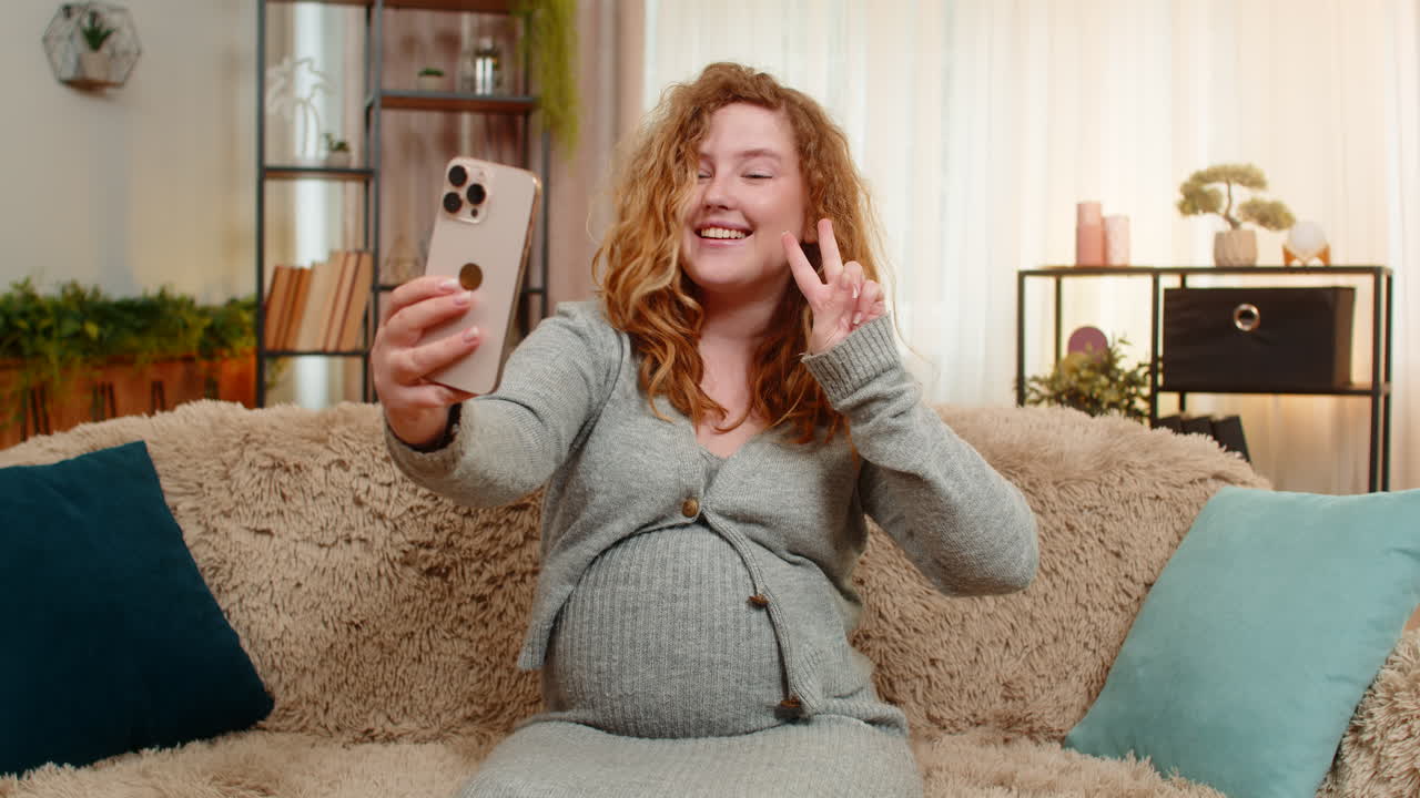 Pregnant woman taking selfie with baby bump on smartphone smiling gently touching belly at home