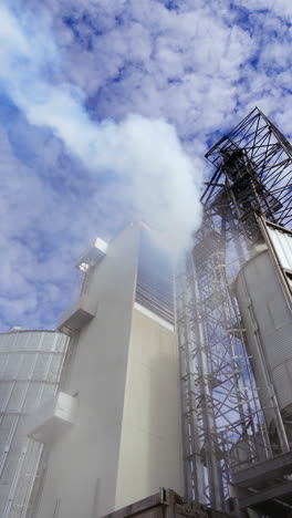 modern plant with metal elevator for agribusiness. white smoke from industrial grain dryer on sky background. storage and further processing of grain. view from below. vertical video