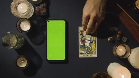Draufsicht-Einer-Person,-Die-Mit-Einem-Mobiltelefon-Mit-Grünem-Bildschirm-Neben-Einer-Sterbekarte-Auf-Dem-Tisch-Eine-Tarotkarte-Liest