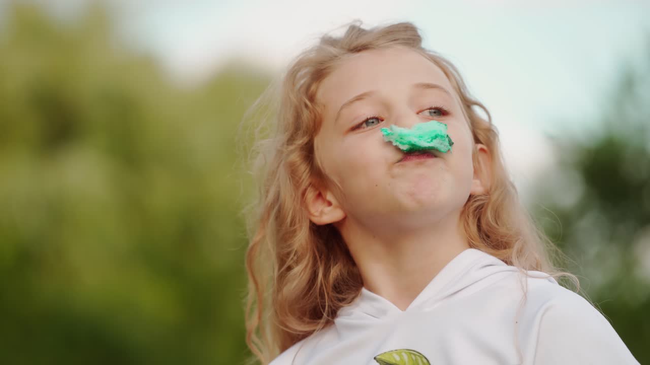 Funny girl with a piece of sweet cotton candy on her face. Pretty little girl eating green cotton candy on blur nature background.