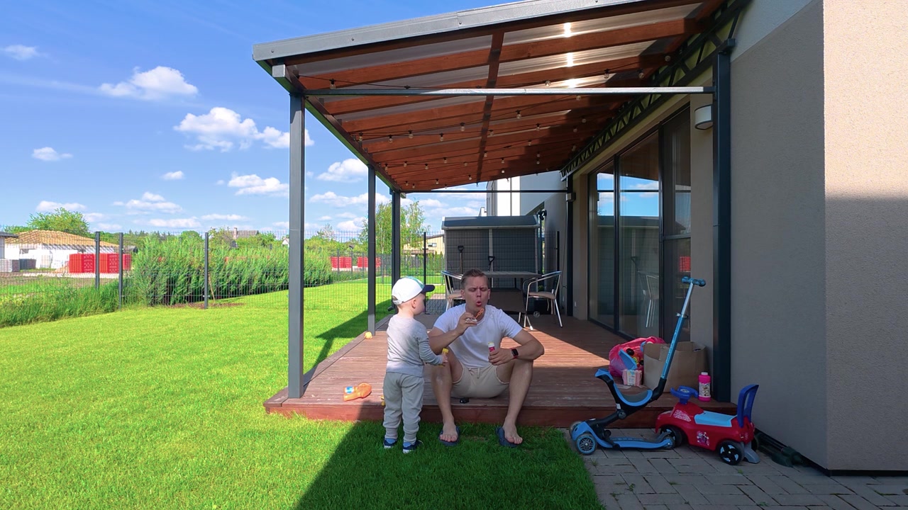 Father and young child spend quality time together on a shaded wooden patio in a modern backyard. Vibrant green grass, sunny weather and relaxed mood create a warm family atmosphere.