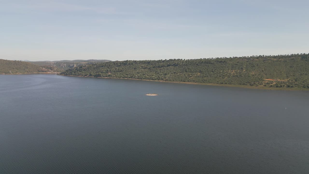 Aerial descending view of Cíjara Reservoir in Badajoz, Spain, highlighting a small island surrounded by calm blue waters under a clear sky. A tranquil natural summer landscape of serenity and beauty