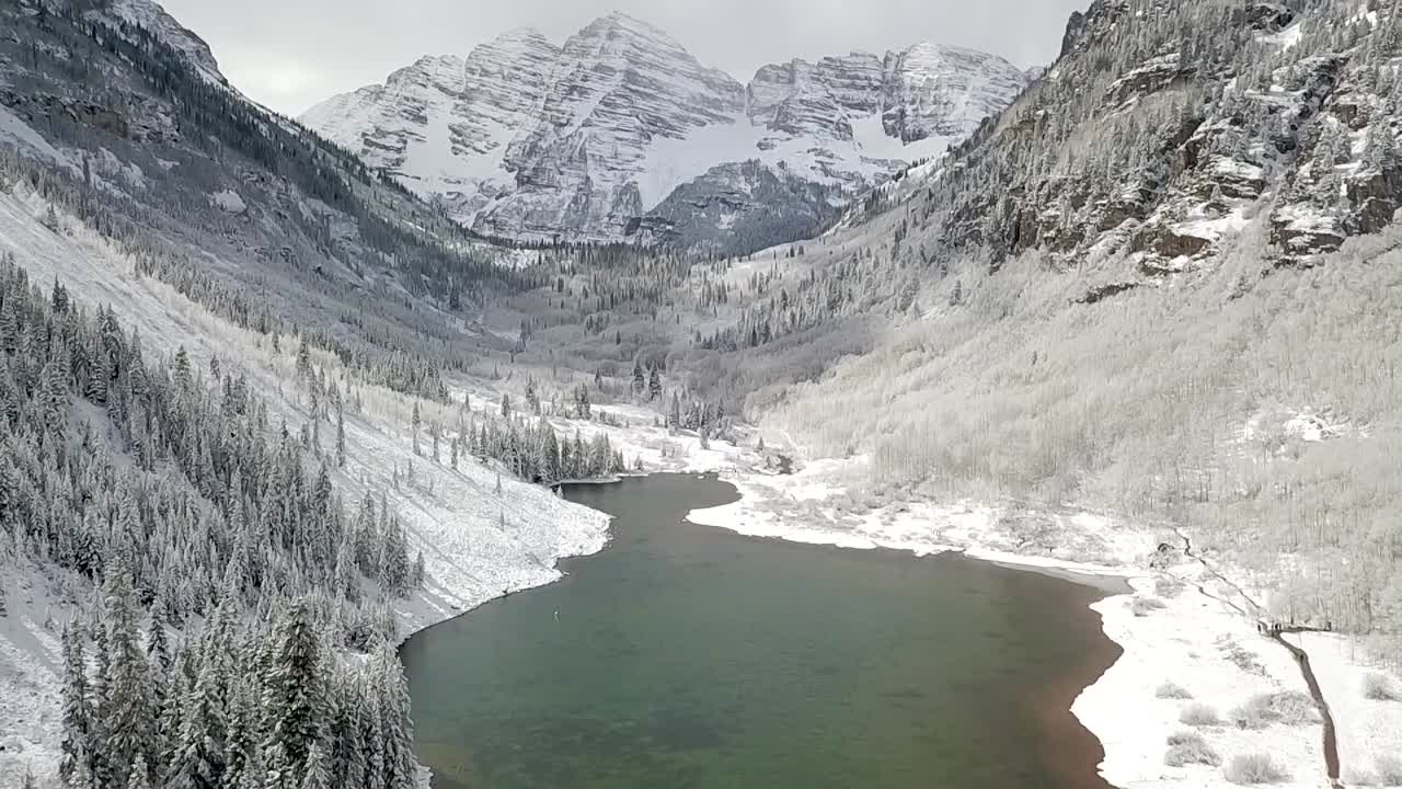 Tilting aerial shot of a dark green lake in a valley of the Maroon Bells Aspen mountain range during the winter season