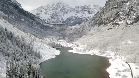 tilting aerial shot of a dark green lake in a valley of the maroon bells aspen mountain range during the winter season