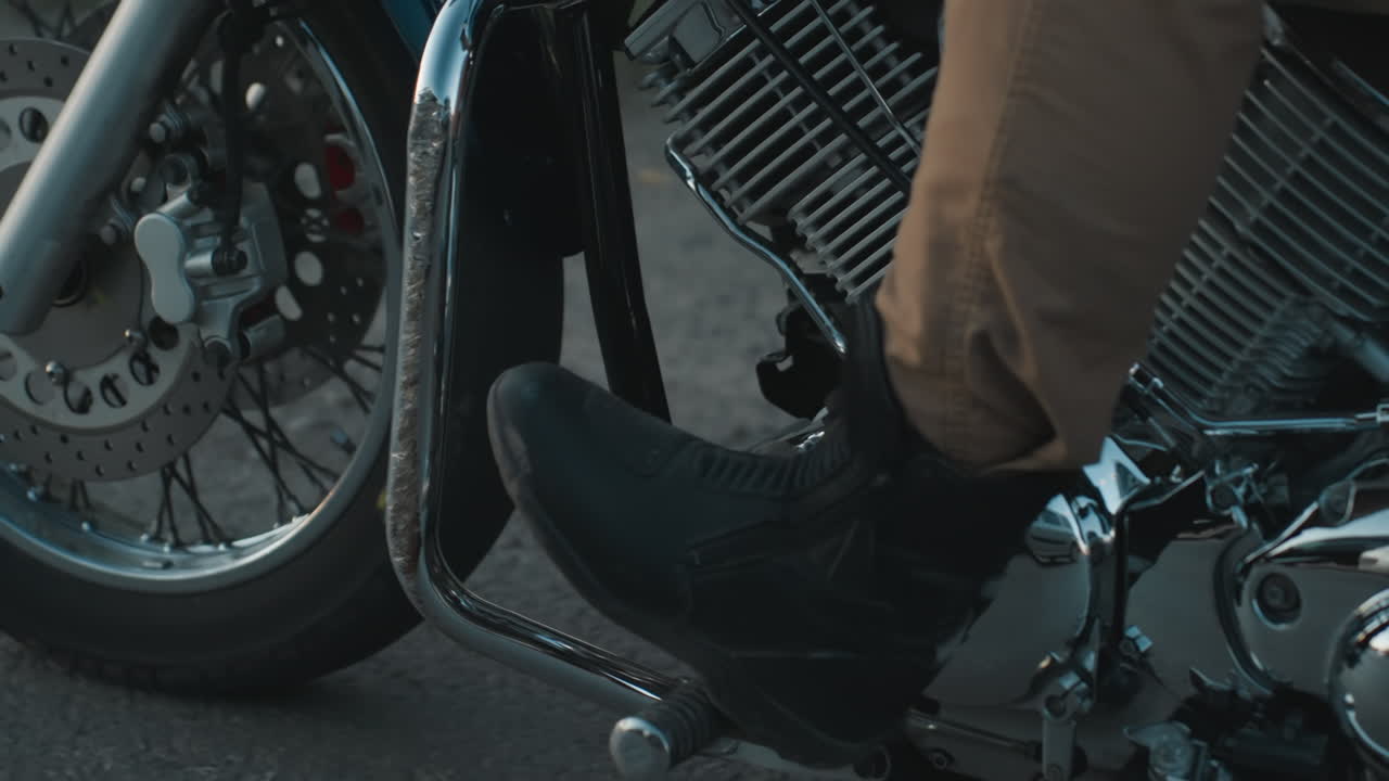 Primo piano della ruota di una motocicletta e dettaglio cromato del motore mentre un motociclista si ferma sulla strada a causa del traffico, mostrando il disco del freno, il pneumatico e le parti metalliche lucide con lo stivale visibile nell'inquadratura