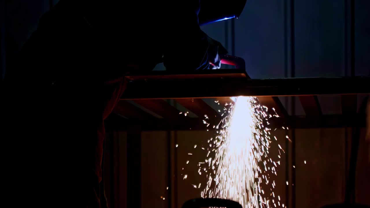Industrial worker welding metal frame with bright sparks in dark workshop