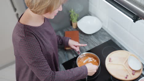 vista aérea de una mujer cocinando la cena en la estufa, agitando la comida en la olla mientras se centra en la preparación de la comida, configuración de cocina moderna con bandeja de madera, cuencos blancos y planta en olla
