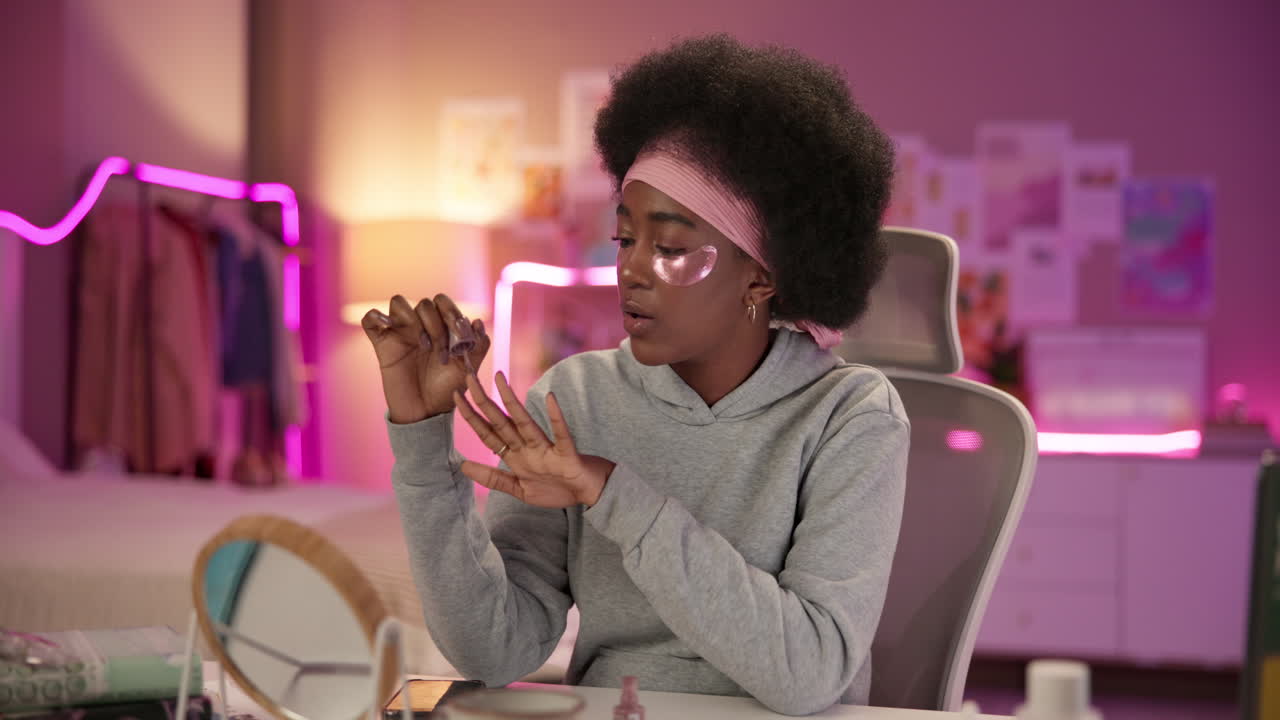 Young African American woman applying nail polish while sitting in a chair in her bedroom