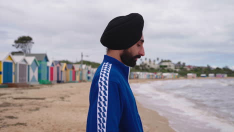 side view of indian man in turban looking and thinking while standing on the dendy street beach with brighton bathing boxes in australia