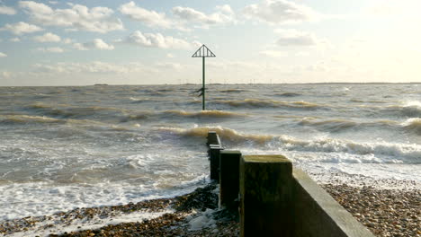 Waves-crashing-groynes-on-the-shore-of-the-beach-with-a-rough-sea-at-sunset-on-a-windy-day,-slow-motion-shot