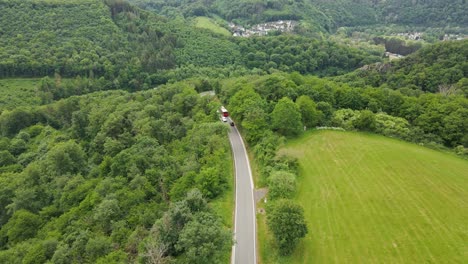 A-red-bus-and-three-cars-travelling-along-a-beautiful-road-leading-down-a-steep-hill-in-between-lush-meadows-and-deciduous-forests-on-a-sunny-spring-morning