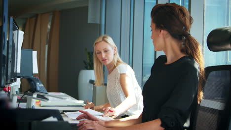 Two-Businesswomen-Working-At-Desks-Have-Discussion-Together