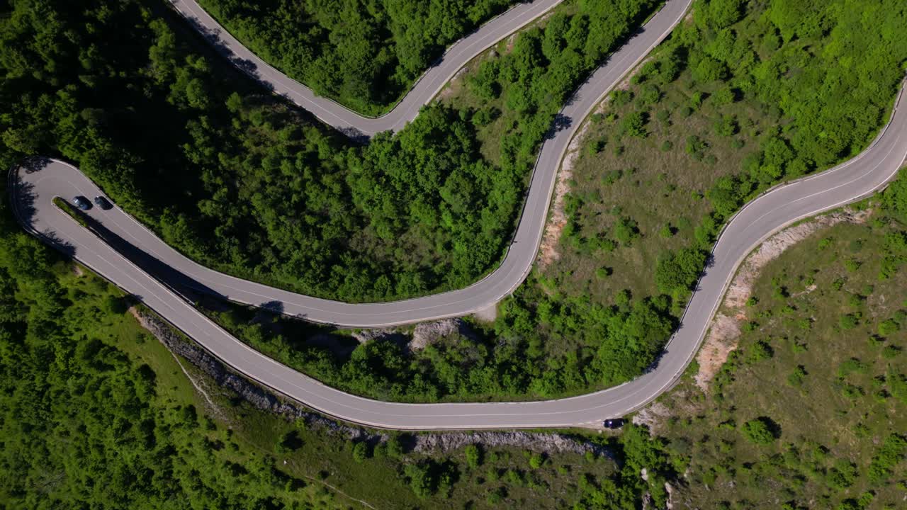 Overhead drone shot of a winding mountain road with sharp hairpin curves cutting through green hills. Shot at Selvino, Italy (Selvino, Italia)