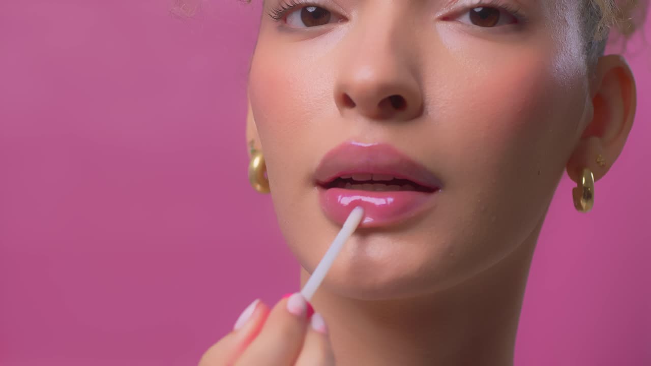 Close up of woman applying pink lip gloss with applicator on a pink background