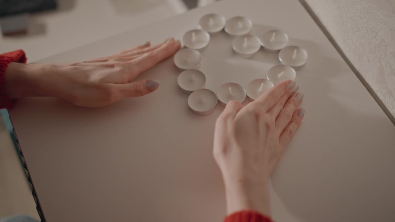 Pair of hands arranging unlit round tealight candles into symmetrical circular ring on off-white tabletop; palms and fingertips guide and smooth placement with subtle hovering and motion blur