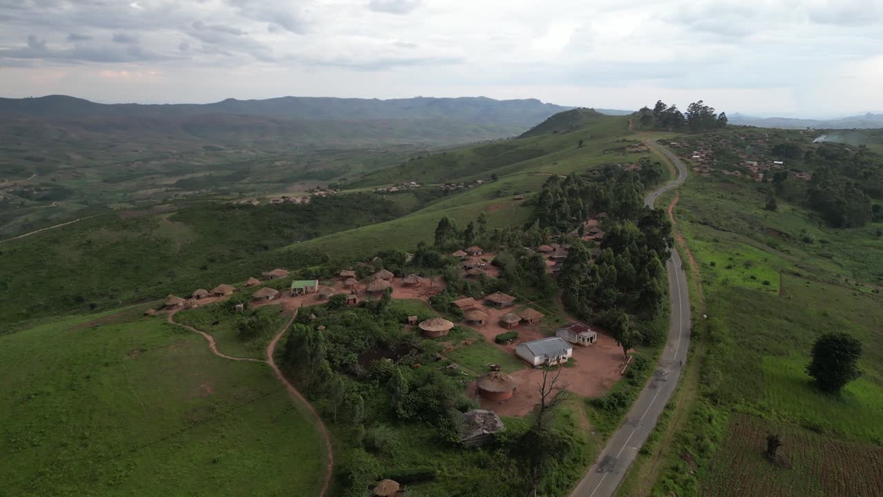 Drone captures traditional mountaintop village with round huts, green hills, and curving road. Scattered homes blend into natural terrain with dramatic views of rolling countryside and cloudy sky
