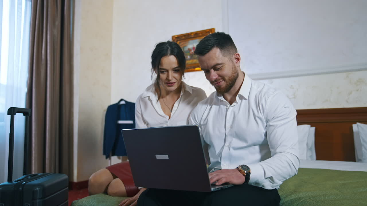 Young couple on trip wedding. Happy guy and girl sitting together on a bed and looking into the laptop in a hotel room. Man and woman at business trip.