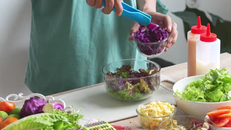 a man making salad, putting purple cabbage sliced into salad bowl, preparing healthy vegan food at home