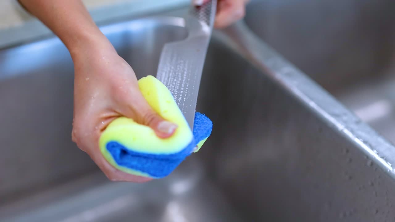 A person cleans a knife with a sponge in a kitchen sink, highlighting effective cleaning techniques and safety