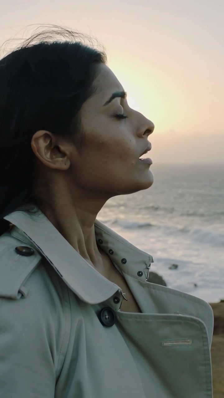 Video verticale: Una donna che inspira si gira verso il sole su una scogliera, la brezza le solleva i capelli, il cappotto e la collana si muovono.