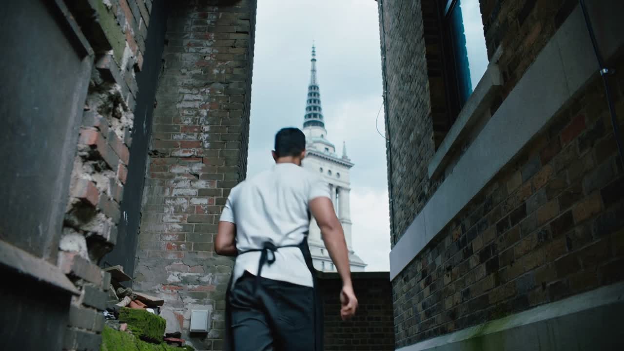 Con un sacco della spazzatura in mano, un operaio si prepara e poi cammina attraverso un vicolo di mattoni verso una torre per smaltire i rifiuti.