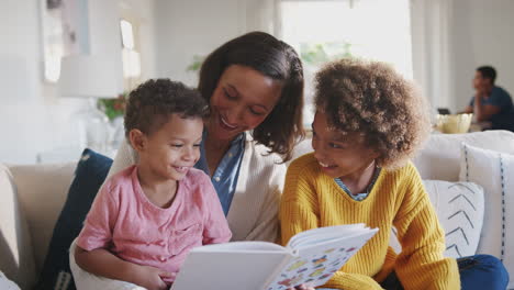 joven madre afroamericana leyendo un libro con sus dos hijos, padre sentado en la mesa en el fondo