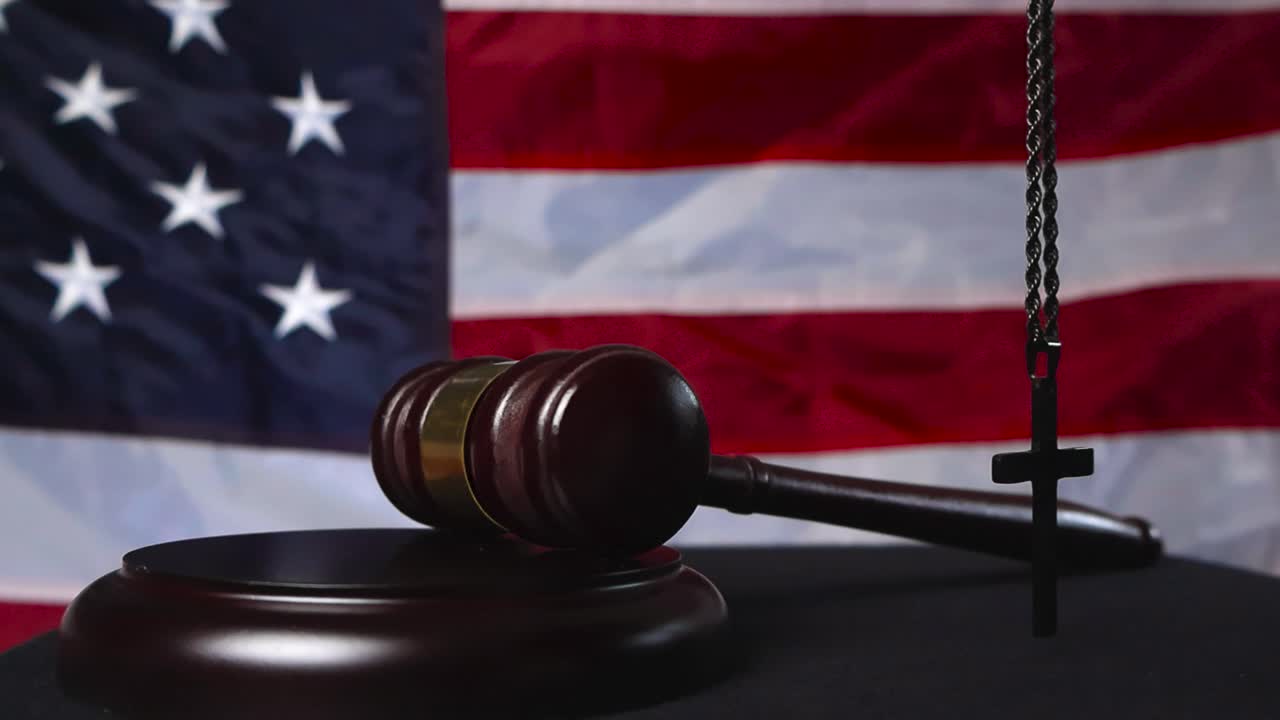 Wooden brown court hammer slammed on a wooden pad by a court justice with a holy cross hangin in the foreground and United States of America flag in the bokeh blurry background. Slow motion movement
