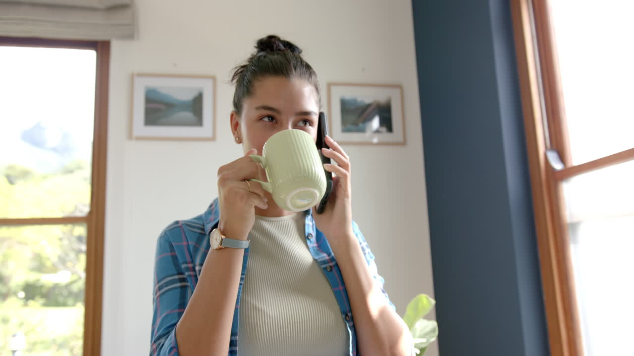 una adolescente biracial feliz bebiendo té, hablando en su teléfono inteligente en casa, copiando espacio, cámara lenta