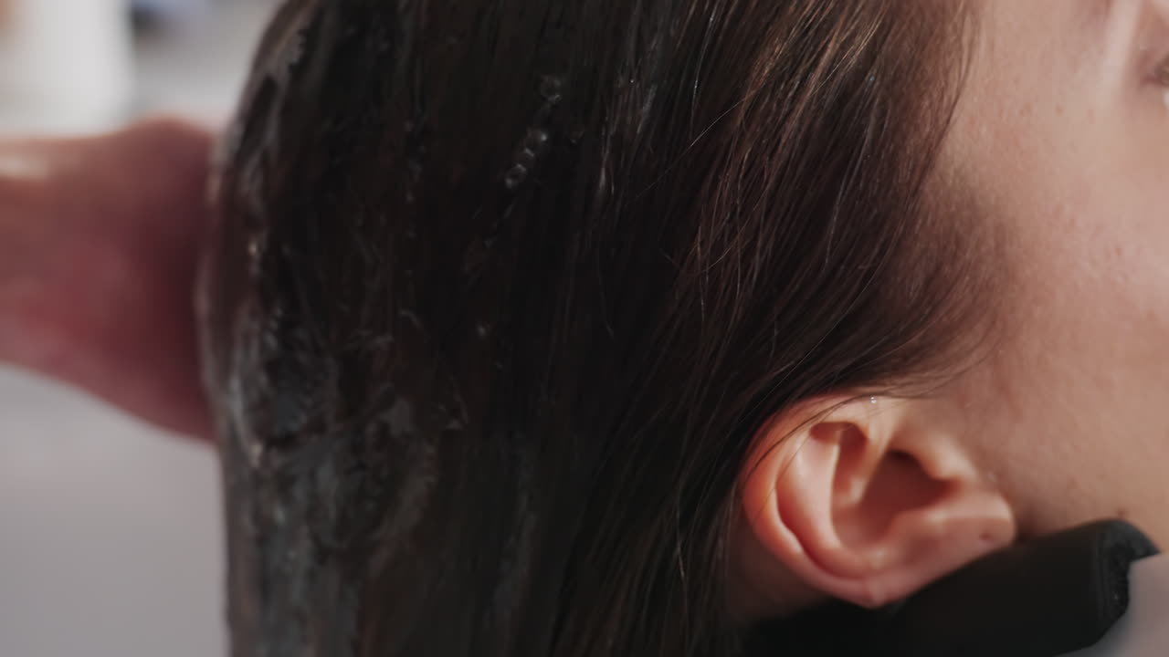 미용실 세면대에서 흐르는 물을 맞고 있는 클로즈업된 백인 여성, 스타일리스트가 머리카락에 따뜻한 물을 붓고 있고, 젖은 머리카락과 물방울이 보이며, 차분한 표정, 전문적인 샴푸 환경, 친밀한 스파 분위기