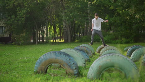 a young boy in a white shirt and gray pants runs across the first two rows of tires in a grassy field, captured in slow motion as he pauses on the last tire in the middle row