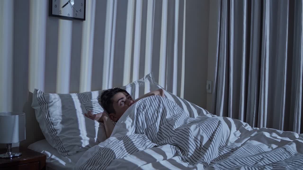 Un'inquadratura video da un'angolazione bassa di una persona a letto, illuminata da ombre a strisce create dalle tapparelle
