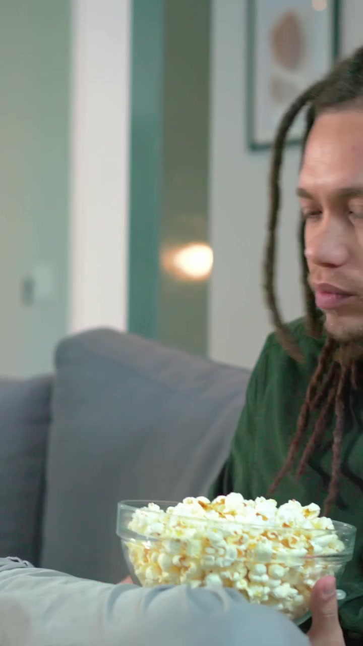 Video verticale: Uomo in camicia verde apre gli occhi, reagisce a qualcosa fuori campo e prende popcorn sul divano di casa.