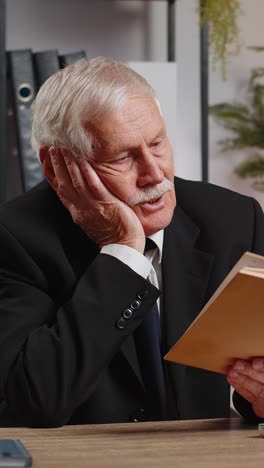 Bored caucasian senior businessman reading book literature taking a rest after work at table