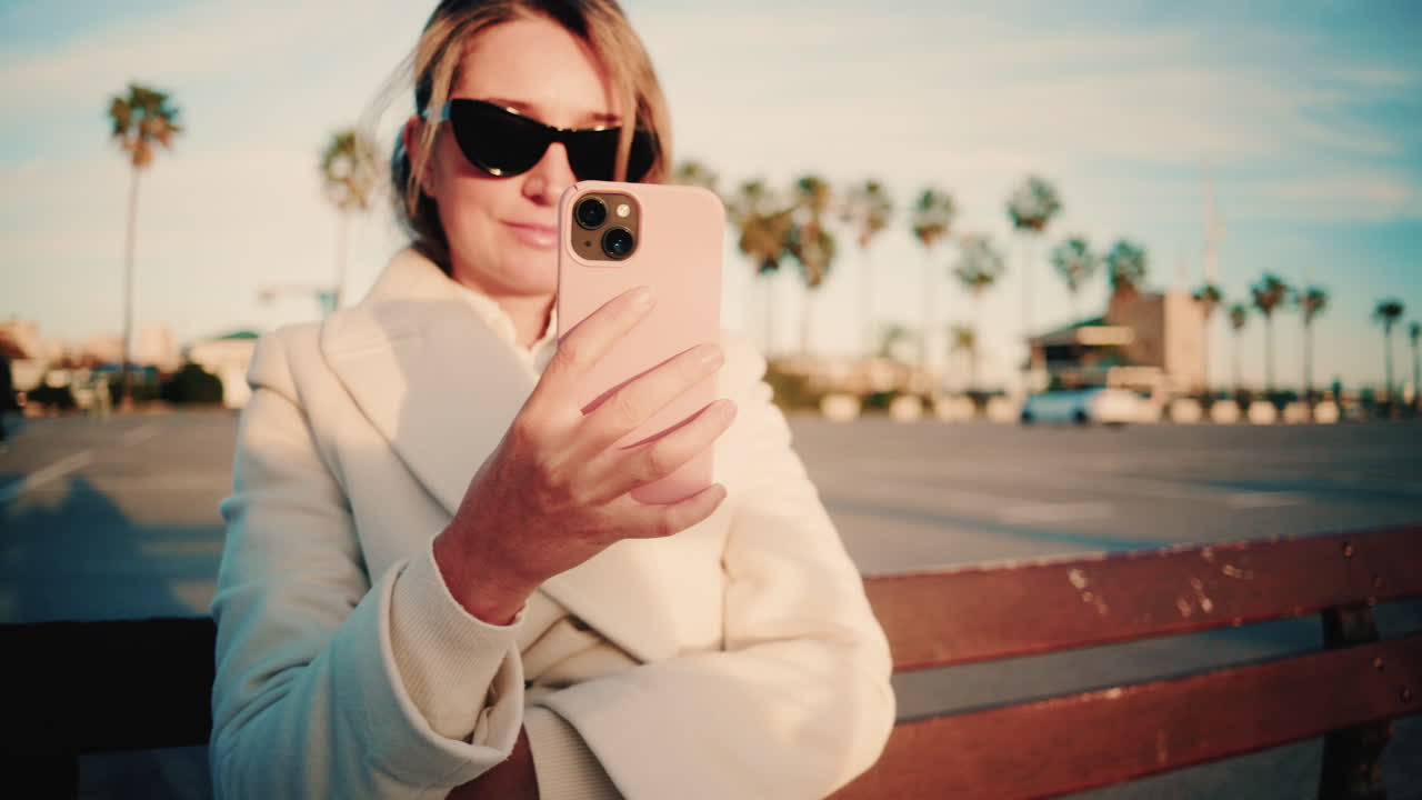 Donna elegante con cappotto bianco guarda il suo smartphone mentre è seduta su una panchina di legno in un parcheggio soleggiato costeggiato da palme