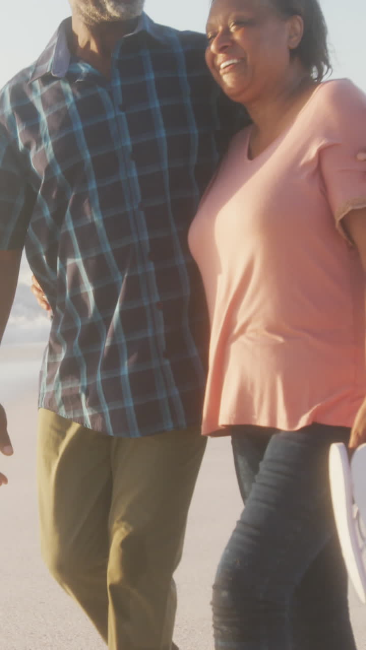 una pareja afroamericana mayor sonriente abrazándose y caminando por una playa soleada