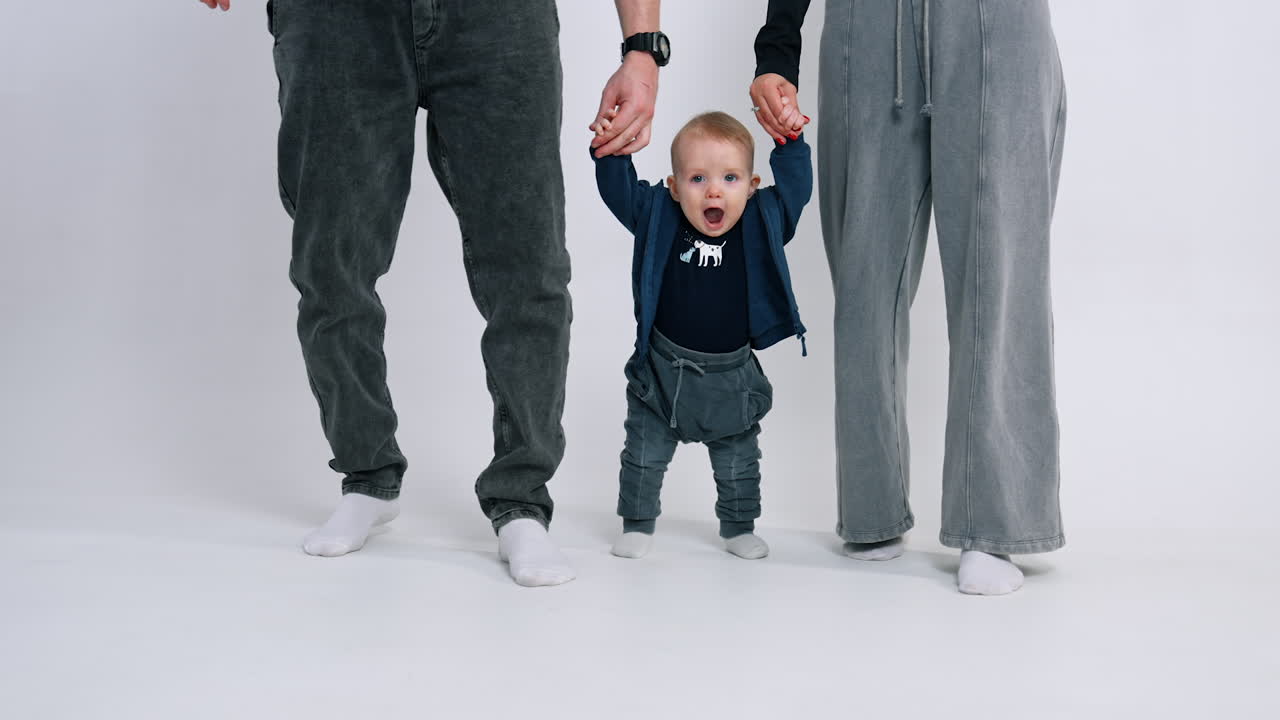 Small baby boy making steps supported by his parents. Family of three wearing jeans. White backdrop.