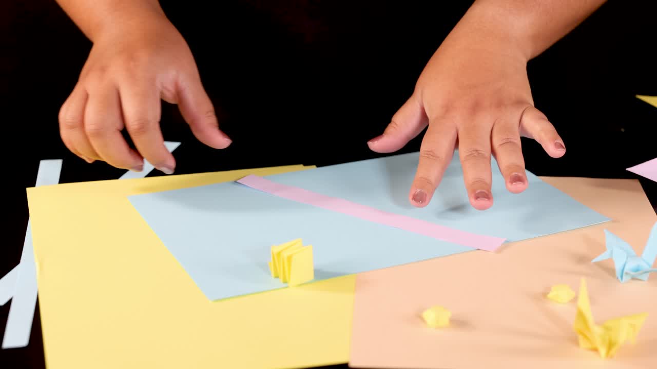 Person folds pink paper strip for origami, overhead view, bright lighting, colorful craft workspace