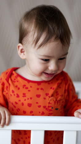 adorable smiling one year old boy stands in his crib laughing cheerfully. lovely baby looks down holding by the handrail. vertical video