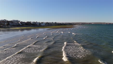 small waves rolling gently on the beach of puerto madryn in chubut, argentina, create a tranquil atmosphere on a sunny day, under a bright blue sky and calm sea, drone descending dolly in shot
