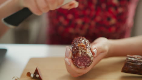 food preparer hands peel tough casing from cured pepperoni end using chef knife on wooden board, revealing rich marbled interior under soft window light in modern kitchen prep scene