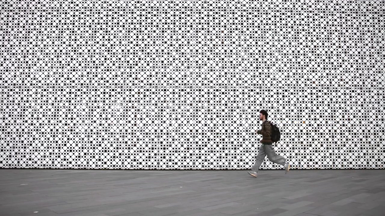 Eén voetganger loopt over een grijs geplaveid plein naast een grote witte geperforeerde metalen gevel, gevormd uit modulaire vierkante panelen met een dicht cirkelvormig gatenpatroon, wat een geometrisch ritme en contrast creëert.
