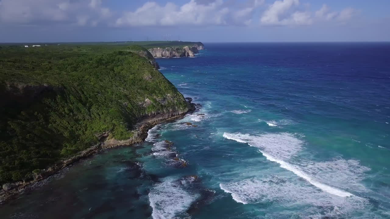 공중에서 바다에서 바위가 많은 녹색 바다 절벽까지 180도 패닝, 바람 터빈이 있는 꼭대기에서 야자수가 있는 해변까지