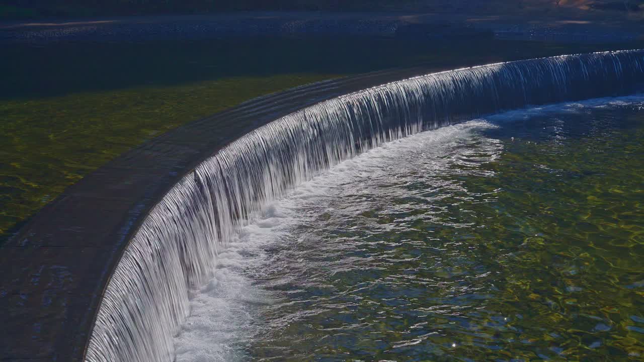 A high-angle shot of water cascading over a curved stone weir into a clear pond. Sunlight reflects on the smooth water surface in a peaceful garden