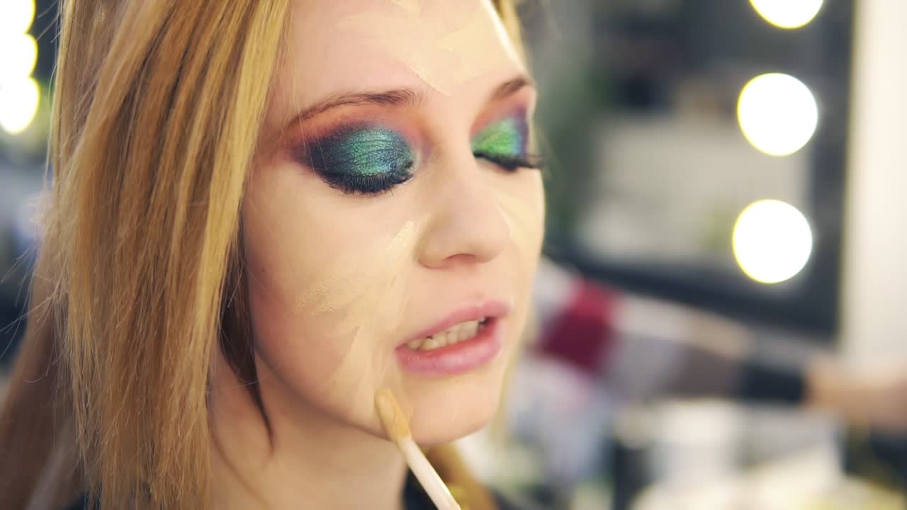 Visagist applying fluid foundation on young smiling woman's face using stick. Professional makeup in salon. Side view