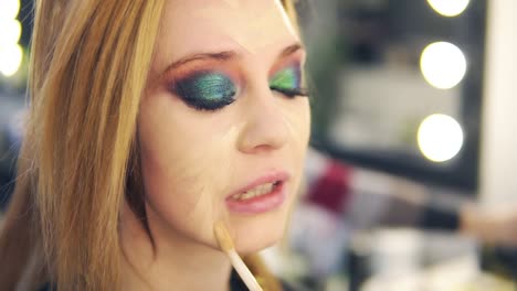 Visagist applying fluid foundation on young smiling woman&#039;s face using stick. Professional makeup in salon. Side view