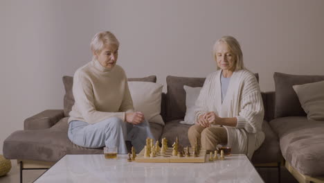 two senior women playing chess sitting on sofa at home 1