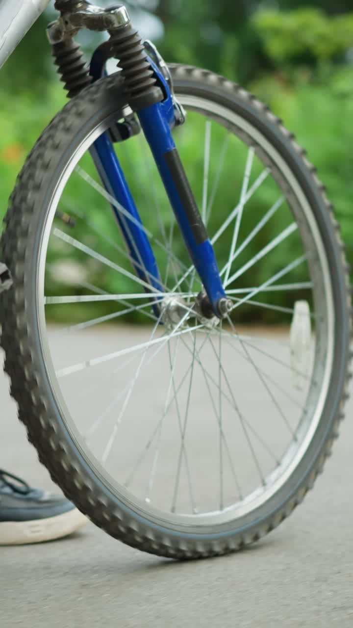 vista de la pierna de alguien caminando de cerca con su bicicleta a lo largo de una pasarela del parque, con cordones de zapatos flotantes en movimiento, el enfoque está en las piernas y la bicicleta, con un fondo de hierba y vegetación